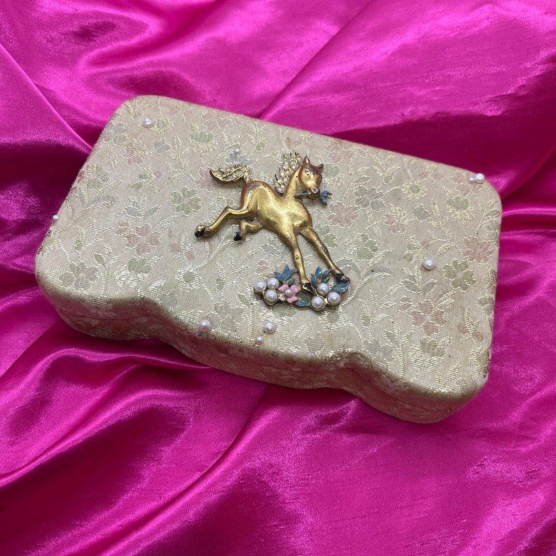 May include: A vintage trinket box with a gold horse design and pearl accents. The box is covered in a floral patterned fabric and has a slightly curved shape.