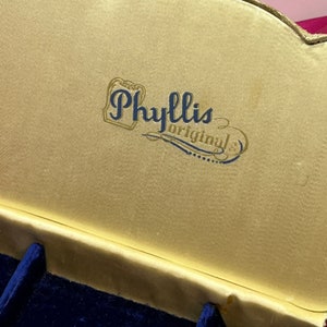 May include: A gold-colored box with a blue velvet interior. The box has a gold label with the text "Phyllis Originals" in blue lettering.