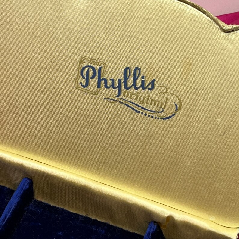 May include: A gold-colored box with a blue velvet interior. The box has a gold label with the text "Phyllis Originals" in blue lettering.