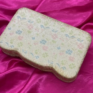 May include: A cream-colored, floral patterned jewelry box with a slightly curved shape. The box is made of a fabric-like material and has a delicate floral design in shades of pink, blue, and green.