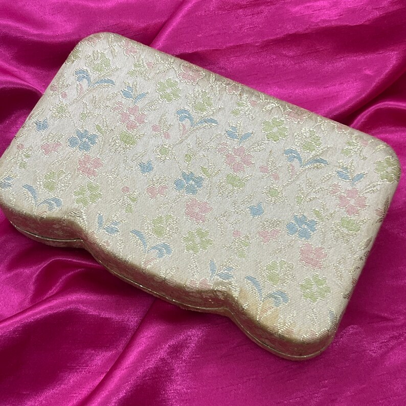 May include: A cream-colored, floral patterned jewelry box with a slightly curved shape. The box is made of a fabric-like material and has a delicate floral design in shades of pink, blue, and green.