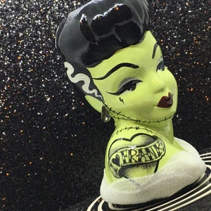 May include: Green ceramic figurine of a woman with black hair, wearing a white bandage and a heart tattoo that says "I Love Frank".