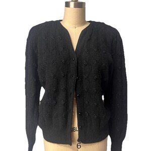 May include: A black long-sleeve cardigan featuring a V-neckline, a front button closure, and a textured knit pattern decorated with small, evenly spaced black beads. The garment has ribbed cuffs and a ribbed hem, providing a structured fit.