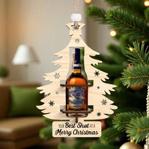 Puede incluir: Adorno navideño de madera con forma de árbol, que contiene una botella de licor. El adorno tiene una estrella en la parte superior y el texto "YOUR Best Shot at a Merry Christmas" en la parte inferior. Cuelga de un árbol de Navidad.