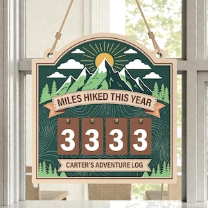 May include: Wooden sign with a mountain and sun design, featuring the text "MILES HIKED THIS YEAR" and a counter showing "3333". The sign includes the text "CARTER'S ADVENTURE LOG". It is hanging by a rope.