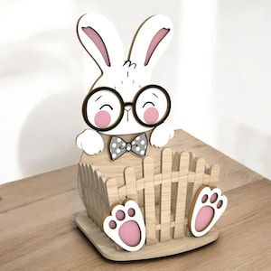 May include: A wooden Easter bunny decoration with a white body, pink inner ears, and a smiling face wearing round black glasses. The bunny has a gray polka-dot bow tie and is perched above a wooden fence and paw-shaped feet.