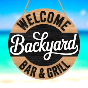 Puede incluir: Cartel de madera redondo con las palabras "WELCOME Backyard BAR & GRILL" en blanco y negro. El cartel está colgado con una cuerda sobre un fondo de playa borroso.