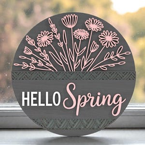 May include: A round, grey wooden sign featuring pink floral cutouts and the words "HELLO Spring". The sign has a decorative border and is designed for spring decor. The sign is approximately 25 cm in diameter.