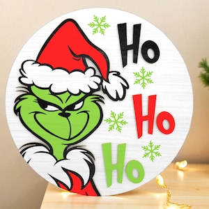 May include: Round wooden Christmas sign featuring the Grinch wearing a Santa hat. The sign has the words "Ho Ho Ho" in black, red, and green, with green snowflake accents. The Grinch is green with a red and white hat.