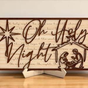 May include: Wooden sign with the words "Oh Holy Night" cut out in a decorative font. The sign features a star, a nativity scene, and the lyrics of the Christmas carol. The sign is displayed on a wooden stand.