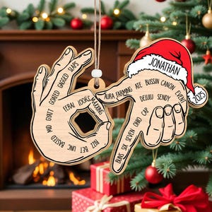 May include: Wooden Christmas ornament shaped like hands making the OK gesture, with text including "Jonathan" and "2025". The ornament is hanging in front of a Christmas tree and fireplace.