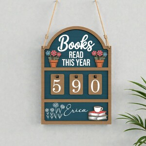 May include: Wooden sign with the text "Books Read This Year" and the number "590". The sign has a teal background with a brown frame and is decorated with flowers and a stack of books with a coffee cup.