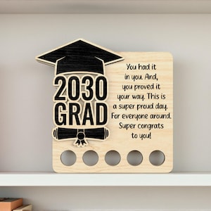 Puede incluir: Placa de graduación de madera con birrete negro, el texto "2030 GRAD" y un diseño de pergamino. La placa incluye el mensaje: "Lo tenías en ti. Y, lo demostraste a tu manera. Este es un día súper orgulloso."