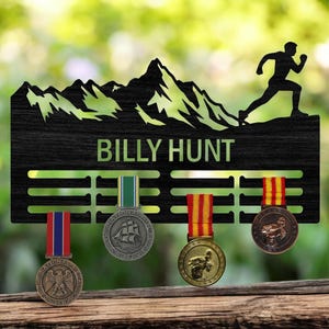 Op de afbeelding: Zwart houten medaille-display met een berg- en hardloper-silhouet. De naam "BILLY HUNT" staat in het groen. Vier medailles met gekleurde linten hangen eronder. Een decoratief stuk voor het tonen van prestaties.