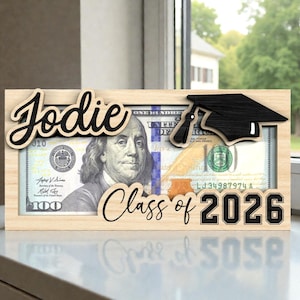 May include: A wooden frame with a $100 bill inside, personalized with the name "Jodie" and "Class of 2026". A black graduation cap is on the right side of the frame. The frame is sitting on a white surface.