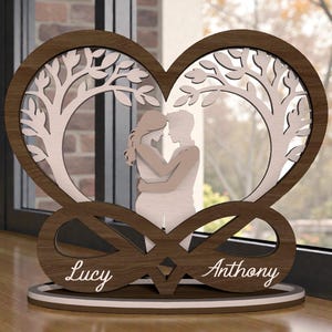 May include: A wooden heart-shaped ornament featuring a tree design and a couple embracing. The names "Lucy" and "Anthony" are inscribed. The piece is brown, white, and beige, with an infinity symbol at the base.
