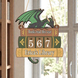 May include: Wooden sign featuring a green and black dragon perched atop a stack of books. The sign reads "Books read this year," with a counter showing "567" and "Erica's Library" at the bottom. The sign is hanging by a string.