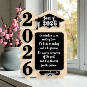 May include: Wooden graduation sign with the year 2026 in large black numbers. The sign includes the text "Class of 2026" and a message about graduation. A vase of pink flowers is in the background.