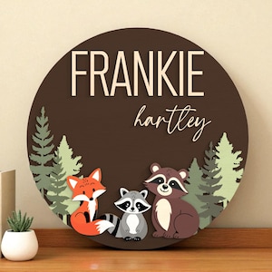 Peut inclure: Panneau rond en bois avec les noms "FRANKIE" et "hartley" en lettres blanches. Le panneau présente un renard, un raton laveur et un ours avec des arbres verts sur un fond marron. Un objet décoratif pour une chambre d'enfant.