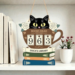 May include: Wooden sign with a black cat peeking over a brown teacup that reads "BOOKS READ THIS YEAR" with the number "963". Below the teacup are three stacked books and a banner that says "ERICA'S LIBRARY".