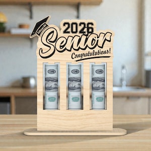 May include: A wooden graduation gift holder with the text "2026 Senior Congratulations!". The holder has three slots, each containing rolled-up US dollar bills. A graduation cap cutout is on top, with the name "Edwards".