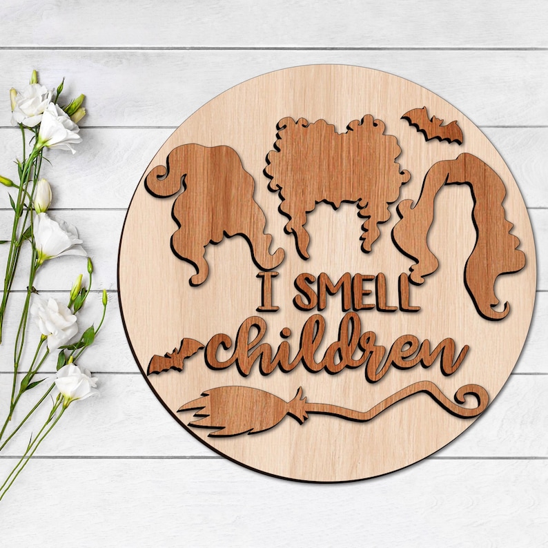 May include: A round wooden sign with a cutout design of three witches with the text "I smell children" and a broom underneath.
