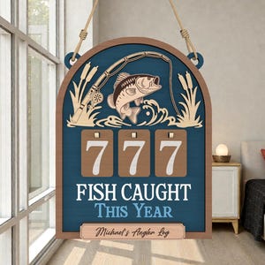 Puede incluir: Cartel de madera con temática de pesca, con fondo azul y marco marrón. El cartel presenta un pez, una caña de pescar y el texto "FISH CAUGHT THIS YEAR". Incluye un espacio para personalizar el cartel con un nombre.