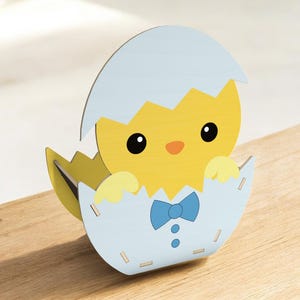 May include: A wooden Easter decoration featuring a yellow chick emerging from a light blue eggshell. The chick has a small orange beak, black eyes, and a blue bow tie. The eggshell is light blue and yellow.