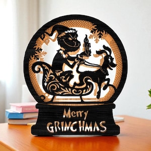 Pode incluir: Decoração de Natal em madeira preta e castanha clara. Apresenta uma silhueta de um personagem num trenó puxado por um cão. O texto "Merry GRINCHMAS" está gravado na base. O design está dentro de uma moldura circular.