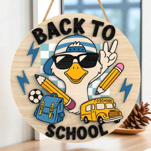 May include: Round wooden sign with the text "BACK TO SCHOOL". The sign features a cartoon duck wearing sunglasses and a hat, holding up a peace sign. Other elements include pencils, a school bus, a backpack, a soccer ball, and lightning bolts.