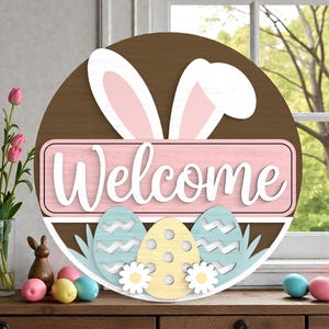 May include: A round wooden Easter sign with a brown background, white bunny ears, and a pink rectangle with the word "Welcome" in white. Below are three decorative eggs in blue, yellow, and white, with grass and flowers.