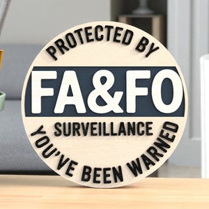 May include: A round wooden sign with the text "PROTECTED BY FA&FO SURVEILLANCE YOU'VE BEEN WARNED" in black and white. The sign has a light wood grain background with a dark blue rectangular section.