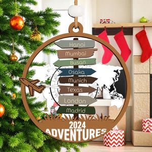 May include: A wooden Christmas ornament with a world map design and directional signs pointing to cities. The ornament features a small airplane and the text "2024 Adventures". The ornament is hanging on a Christmas tree.