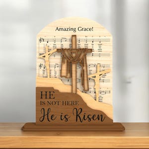 May include: Wooden Easter decoration featuring three crosses, with the central cross draped with fabric. The background includes sheet music and the words "Amazing Grace!" The base reads "He is not here, He is Risen."