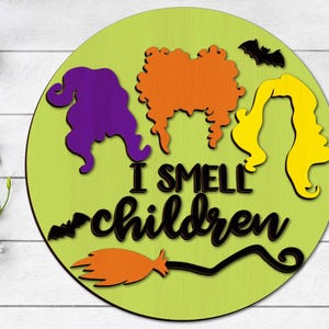 May include: A green round wooden sign with a black silhouette of three witches with the text "I smell children" and a black silhouette of a broom and bat.