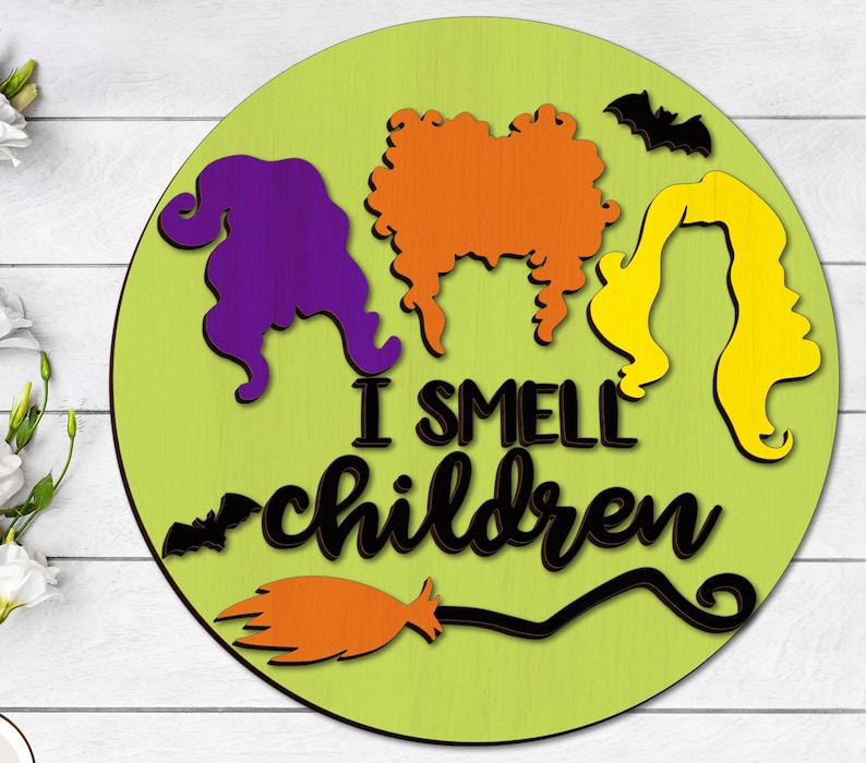 May include: A green round wooden sign with a black silhouette of three witches with the text "I smell children" and a black silhouette of a broom and bat.