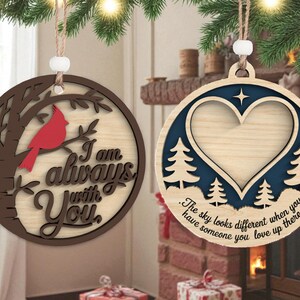 May include: Two wooden Christmas ornaments. One features a red cardinal on a tree branch with the text "I am always with You." The other has a heart-shaped cutout, trees, and the text "The sky looks different when you have someone you love up there."