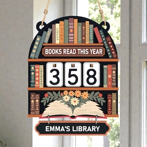 May include: A wooden sign shaped like a bookshelf, with the text "BOOKS READ THIS YEAR" and "358". The sign has a floral and book design, with the text "EMMA'S LIBRARY" at the bottom. It is suspended by a rope.