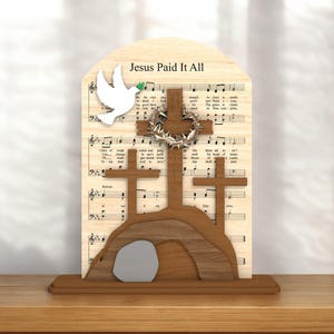 May include: Wooden religious decoration featuring three crosses, a dove, and the text "Jesus Paid It All" on a background of sheet music. The decoration includes a tomb and a crown of thorns.