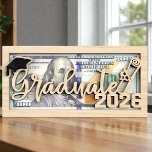 May include: A wooden frame with the word "Graduate" and the year "2026" cut out, with a graduation cap and diploma design. The frame holds a U.S. $100 bill, making it a graduation gift.