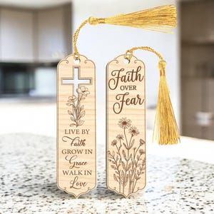 以下が含まれることがあります： 金色のタッセルが付いた2つの木製ブックマーク。1つのブックマークには、十字架と花の模様と「Live by Faith, Grow in Grace, Walk in Love」の文字が描かれています。もう1つのブックマークには「Faith Over Fear」の文字と花の模様があります。