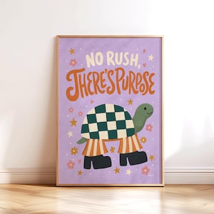 May include: A framed art print featuring a cartoon turtle with a checkered shell and striped pants. The print has the text "NO RUSH, THERE'S PURPOSE" in orange and white lettering on a lavender background, with floral and star accents.