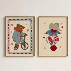 Puede incluir: Dos láminas decorativas enmarcadas con motivos de circo. La lámina de la izquierda muestra un oso pardo con traje de payaso azul montando en bicicleta. La de la derecha muestra un elefante gris con chaqueta azul y pantalón a rayas rojas sobre una bola.