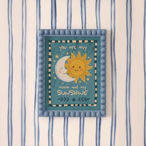 Puede incluir: Una impresión enmarcada con un borde azul con cuentas. La obra de arte presenta un sol y una luna sonrientes con el texto "you are my moon and my sunshine" sobre un fondo turquesa. El fondo es una pared blanca con rayas azules.