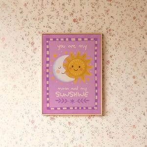 Puede incluir: Un cuadro enmarcado con un sol y una luna sonrientes sobre un fondo lavanda. El texto "you are my moon and my sunshine" se muestra debajo de los cuerpos celestes. El marco es de color marrón claro.