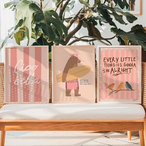 Pu&ograve; includere: Tre stampe artistiche incorniciate con sfondo a righe rosa chiaro e bianche. Una stampa dice "Ciao Bella", un'altra presenta un orso con una tavola da surf e la terza dice "Every Little Thing Is Gonna Be Alright" con uccelli.