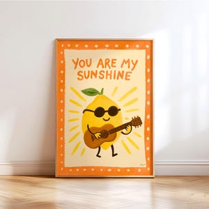 Puede incluir: Una impresión artística enmarcada que presenta un limón de dibujos animados con gafas de sol tocando la guitarra. El limón es amarillo con una hoja verde y está rodeado de rayos de sol amarillos. El texto "YOU ARE MY SUNSHINE" está escrito encima del limón.