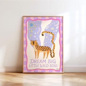 Puede incluir: Un póster enmarcado con un guepardo de dibujos animados con pelaje manchado, frente a un fondo caprichoso con estrellas, lunas y un sol. El texto "DREAM BIG LITTLE WILD SOUL" está en la parte inferior, enmarcado por un borde rosa y morado.