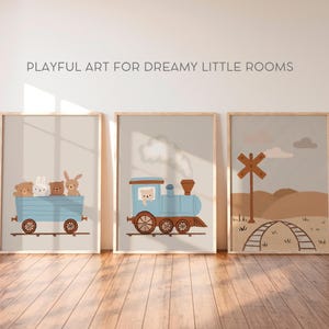 May include: Three framed prints with a whimsical train theme. The first print features a blue train with a brown bear, a white rabbit, and a brown bunny riding in the car. The second print shows a blue train with white smoke coming from the chimney. The third print shows a brown wooden railroad crossing sign with a brown wooden fence and a brown dirt path.