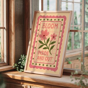 May include: A framed art print featuring the words "BLOOM INSIDE AND OUT" with pink flowers and green leaves. The artwork has a light yellow background with a pink border and star accents. The frame is a light wood color.